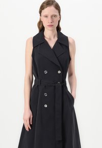 Μαύρο αμάνικο παλτό φόρεμα με μεγάλο γιακά, διπλές κουμπότρυπες και ταιριαστό ζώνη. Το ύφασμα είναι λείο με δομημένο σχέδιο.