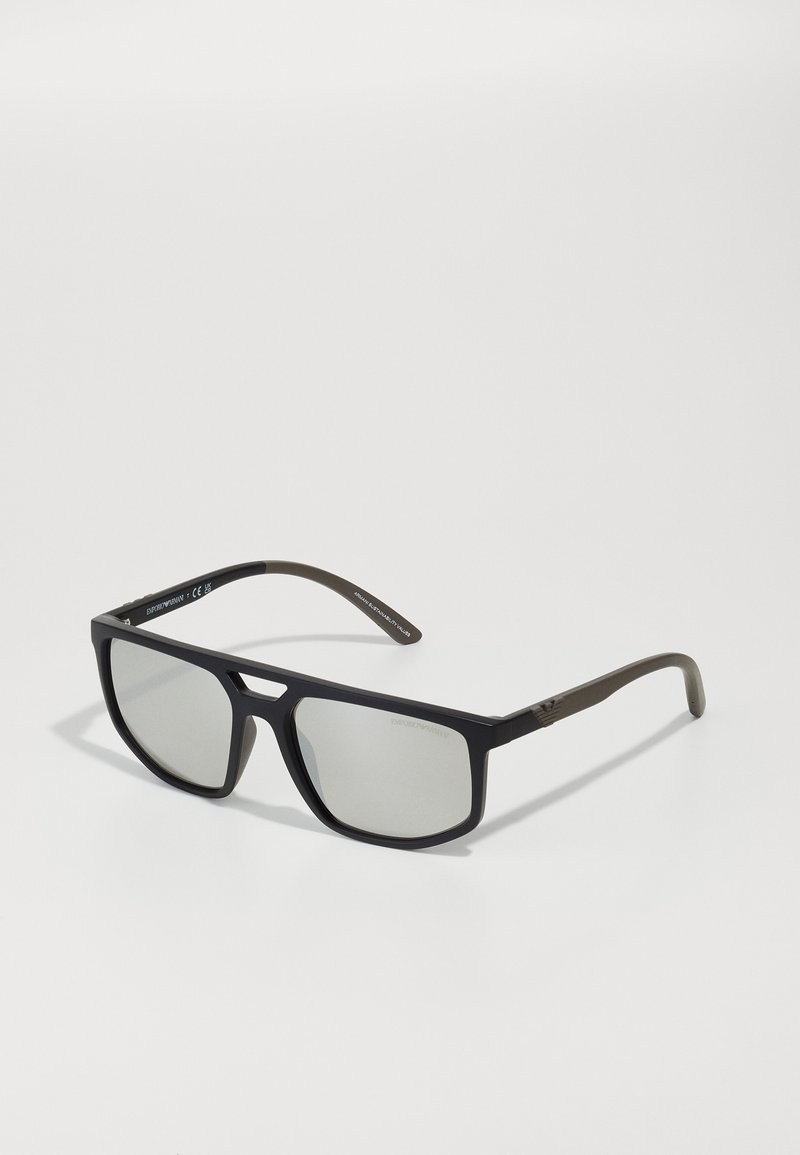 Saulesbrilles ar melnu matētu rāmi, pelēkām tonētiem stikliem un olīvzaļām tempļu rokām. Eleganta ģeometriska dizaina ar logotipa detalizāciju uz tempļiem.