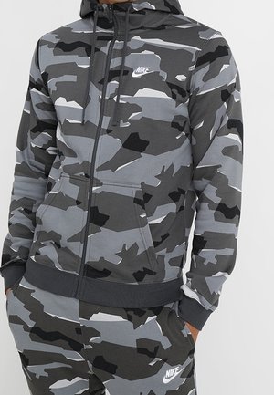 Homme portant un hoodie Nike camouflage gris et noir et un pantalon de survêtement assorti, les mains dans les poches.