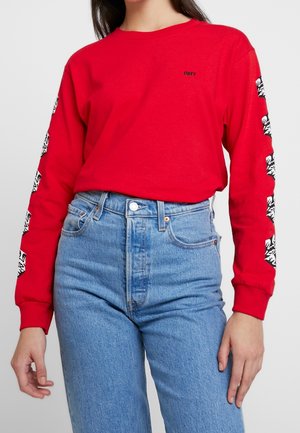Persoon draagt een rood shirt met lange mouwen met zwart-witte grafische strepen op de mouwen en een klein "OBEY"-logo, gecombineerd met een blauwe jeans met hoge taille.