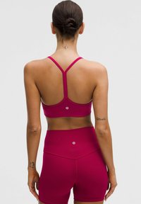 Žena s účesom v bunke, oblečená v burgundskej športovej podprsenke a šortkách s vysokým pásom, zobrazená zozadu s malými tetovaniami na krku a rukách.