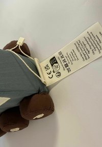 Pliušinis žaislas su pilku kūnu ir rudais apvaliais kojomis, turintis grietinėlės spalvos akcentus ir prisegtą priežiūros etiketes su virvele.