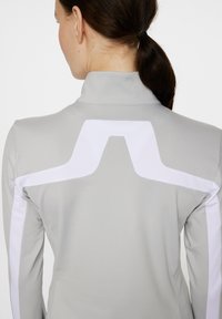 Hellgraues Sportoberteil mit hohem Kragen, das ein weißes geometrisches Design über den Rücken und an den Seiten aufweist. Glattes, dehnbares Material.