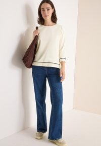 Jasnożółta bluzka z długim rękawem i czarnymi wykończeniami, zestawiona z niebieskimi dzwonami. Modelka trzyma brązową zamszową torbę na ramię. Podłoga jest jasna.