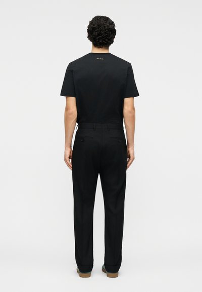 Άνδρας που στέκεται πλάτη με μαύρο κοντομάνικο πουκάμισο με μικρό χρυσό λογότυπο στον λαιμό και μαύρο ραμμένο παντελόνι με καφέ παπούτσια.