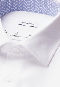 Witte overhemd met een blauw patroon aan de binnenzijde van de kraag, merkembleem "seidensticker," en knopen zichtbaar aan de voorkant.