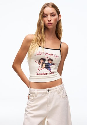 Fiatal nő hosszú szőke hajjal, fehér pántos, csillagos grafikával és szöveggel ellátott crop topban, világos színű nadrággal párosítva.