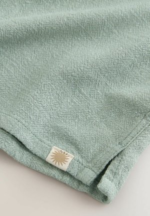 Material texturat verde cu un model geometric subtil, având o etichetă cusută cu un design de soare la tiv. Material moale și țesut.