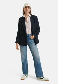 Marineblaues doppelt knöpfbares Sakko, weißes gestreiftes Hemd, hellblaue Jeans und weiße Sneakers mit schwarzen Akzenten, gekrönt mit einer hellen Kappe.
