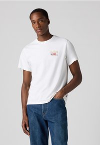 Witte katoenen t-shirt met korte mouwen en ronde hals. Bevat multikleurige gedrukte tekst op de linkerborst. Draagt samen met blauwe spijkerbroek.