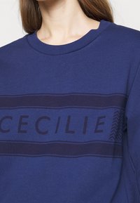 Námořnická modrá mikina s kulatým výstřihem, tmavý pás přes hruď se slovem "CECILIE" a akcentem v podobě cik-cak vzoru.