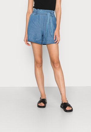 Kvinde iført højtaljede blå denimshorts med papirpose-talje og bundet bælte, sort ærmeløs top og sorte sandaler.