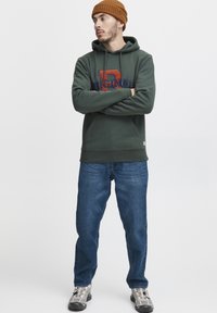 Haki zaļa kapuce ar lielu izšūtu "D" un "ORIGINAL" tumši zilā un oranžā krāsā, saskaņota ar zilām džinsām un pelēkām kedām. Silta adīta cepure.