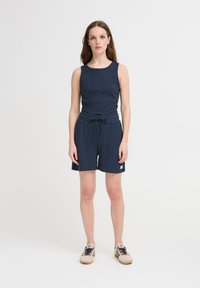 Jumpsuit blu navy a coste con top senza maniche, vita elastica e nodo. Presenta pantaloni corti larghi e una piccola etichetta con logo sul lato. Tessuto casual.
