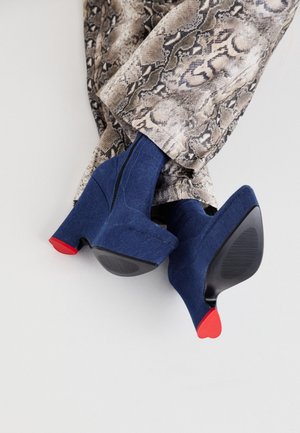 Botines azul ante con plataforma y puntas de tacón rojas, combinados con pantalones de estampado de serpiente sobre un fondo neutro.