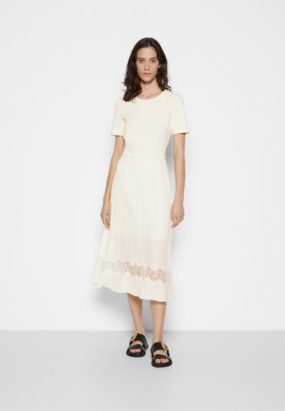 Robe couleur crème avec un haut ajusté et une jupe plissée évasée. L'ourlet inférieur présente des détails en dentelle. Des sandales noires complètent le look.