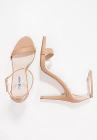 Ein Paar beigefarbene High-Heels-Sandalen mit Knöchelriemen, offenen Zehen und schlanken Absätzen, flach und seitlich auf einem weißen Hintergrund präsentiert.