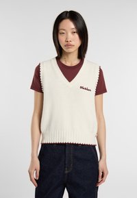 Chaleco de punto color crema con escote en V, ribete acanalado y logotipo burdeos bordado. Llevado sobre una camiseta color marrón y combinado con jeans oscuros.