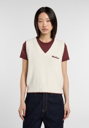 Gilet lavorato a maglia di colore crema con scollo a V, bordi a coste e logo ricamato in bordeaux. Indossato sopra una t-shirt bordeaux e abbinato a jeans scuri.