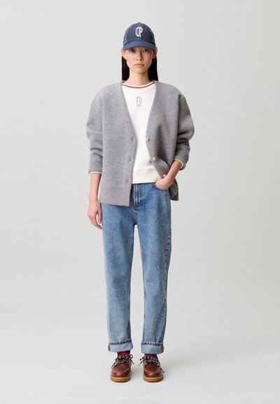 Cárdigan gris oversized, sudadera blanca con ribete rojo, vaqueros azul claro, zapatos marrones y gorra azul con logo blanco.