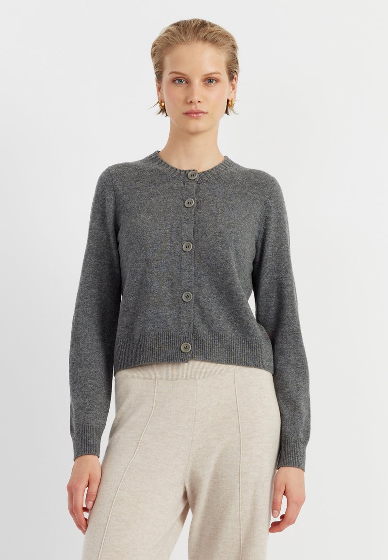 Cardigan grigio lavorato a maglia con scollo rotondo, caratterizzato da sette bottoni sul davanti e polsini e orlo a coste. Indossato con pantaloni beige.