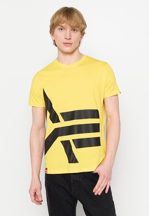 Young man with blond hair wearing a yellow t-shirt with a large black geometric design and black pants standing against a plain background.