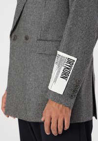 Sacou gri cu model houndstooth, cu design cu două rânduri de nasturi, nasturi maro și etichetă albă de îngrijire pe manșetă.