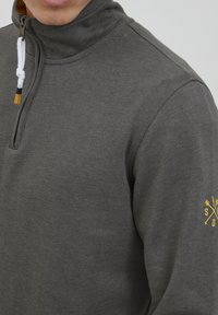 Sudadera gris con cremallera y cuello alto, que presenta un cordón blanco y un detalle amarillo. La manga izquierda tiene un diseño de logo amarillo.