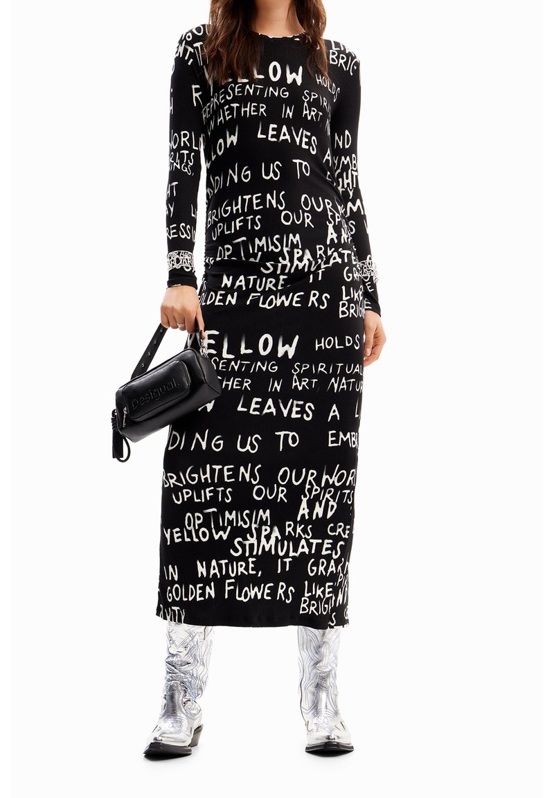 Femme portant une robe noire à manches longues avec un texte manuscrit blanc, tenant un sac à main noir et portant des bottes argentées métalliques.