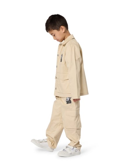 Beżowa bawełniana kurtka z przednimi kieszeniami i haftem, zestawiona z dopasowanymi luźnymi spodniami z naszywkami i grafiką, noszona z białymi tenisówkami.