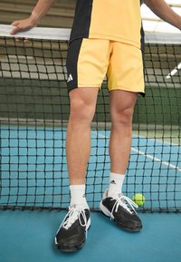 Żółto-czarne szorty sportowe, do kolan, z logo Adidas. Czarne sneakersy o fakturowanej powierzchni i białe skarpetki do kostek, stojące obok siatki tenisowej.