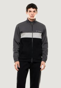 Chaqueta con capucha y cremallera en negro, gris y gris claro. Presenta un diseño a bloques de color, bolsillos frontales y un collar alto. Está hecha de un tejido suave.