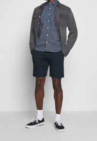 Giacca di denim grigio su una camicia blu a pois, abbinata a pantaloni corti scuri e sneakers nere. Calzini bianchi visibili alle caviglie.