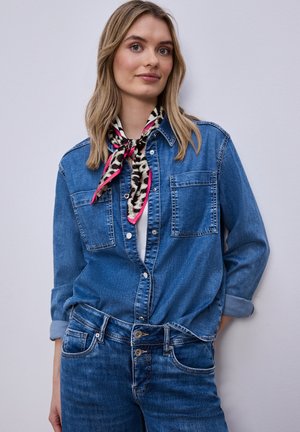 Femme portant une chemise en denim et un jean avec les manches retroussées, accessoirisée d'une écharpe imprimée animal en noir et blanc avec des bords roses.