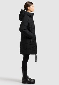 Schwarzer gepolsterter Mantel mit Kapuze, ausgestattet mit einem diamantgequilten Design und seitlichen Taschen. Kombiniert mit schwarzen Knöchelstiefeln und Leggings.