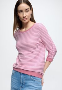 Lichtroze trui met lange mouwen en horizontale geribbelde structuur, ronde halslijn en roze geribbelde manchetten. Gecombineerd met een blauwe spijkerbroek.