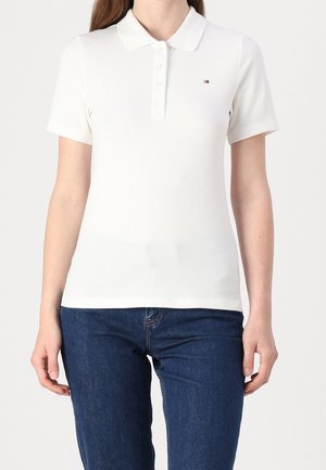 Kobieta ubrana w prostą białą koszulkę polo z krótkim rękawem, zapinaną na guziki z małym logo, w połączeniu z ciemnoniebieskimi dżinsami, stojąca na białym tle.