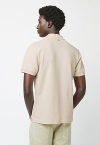 Polo beige con maniche corte, dettaglio dei bottoni al colletto e texture liscia. Indossato con pantaloni verde chiaro.