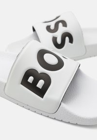 Valkoiset liukusandaalit, joissa on tekstuuripohja, leveä hihna ja rohkea musta "BOSS"-logo painettuna yläosan. Vahva synteettinen materiaali.