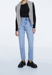 Schwarzes, geripptes Langarmoberteil, weiß-gestreiftes Weste, hellblaue hochgeschnittene Jeans mit Rissen, schwarze Plateauslipper.