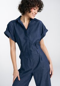 Kombinezon jeansowy w ciemnym niebieskim kolorze, z krótkimi podwiniętymi rękawami, kołnierzykiem, paskiem w talii oraz luźnym krojem z delikatną fakturą.