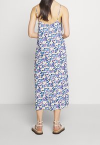 Kwiatowa sukienka maxi w niebieskie, białe i fioletowe wzory; cienkie ramiączka; lekką tkanina; luźny krój; długość do połowy łydki; zestawiona z białymi sandałami.