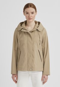 Beige wasserdichte Jacke mit großer Kapuze, Reißverschluss und Seitentaschen. Glatte Textur und lässige Passform, geeignet für verschiedene Wetterbedingungen.