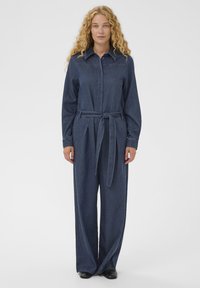 Ženska s kodrastimi blond lasmi, oblečena v temno modro jeans kombinezon s pasom in črnimi čevlji, ki stoji pred enostavno svetlo ozadje.