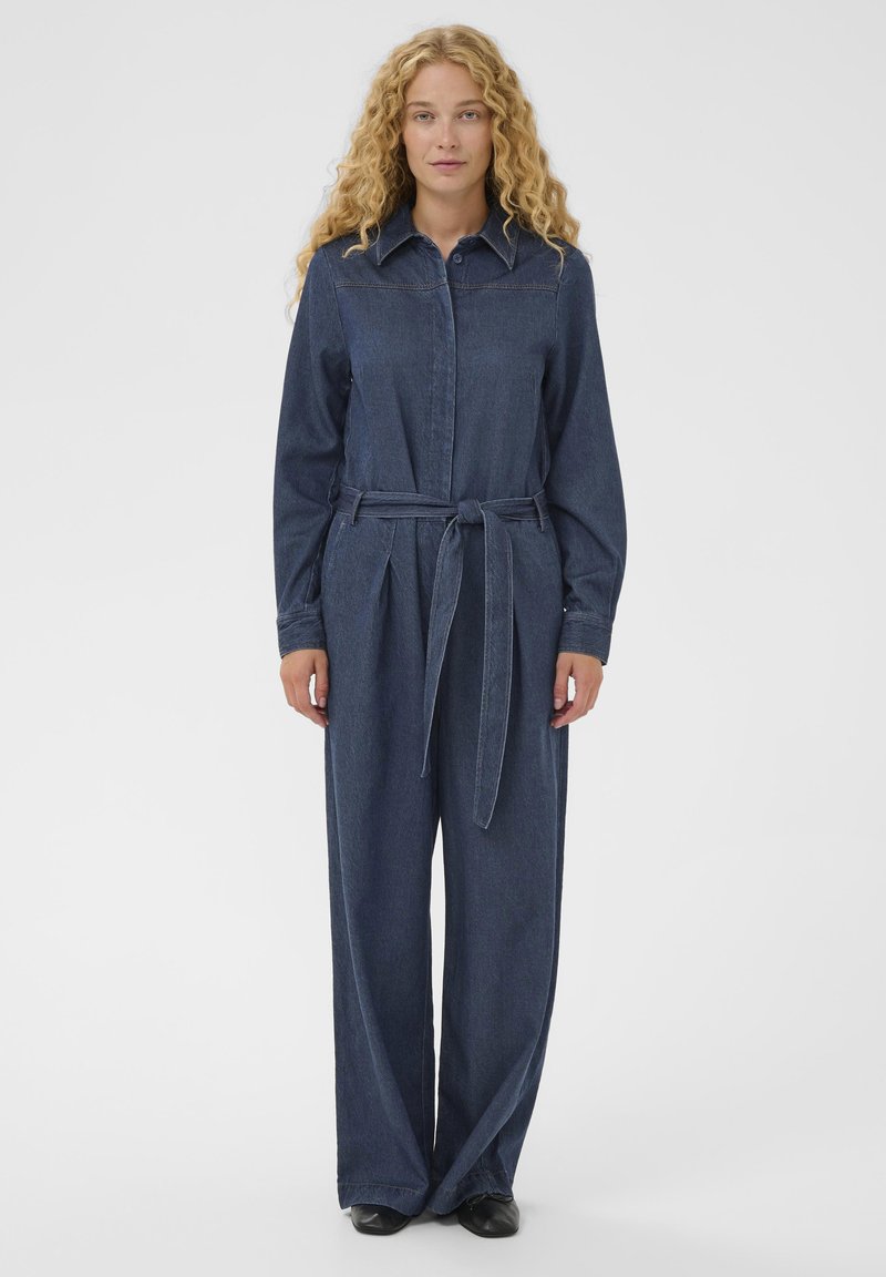 Ženska s kodrastimi blond lasmi, oblečena v temno modro jeans kombinezon s pasom in črnimi čevlji, ki stoji pred enostavno svetlo ozadje.