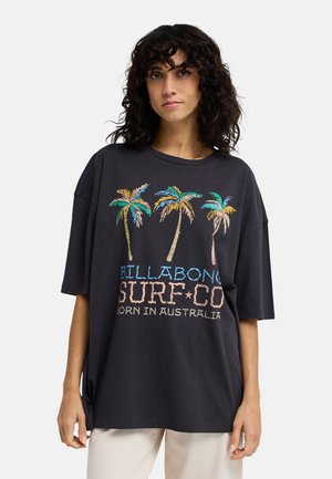 Donna che indossa una t-shirt nera oversize con palme colorate e la scritta «Billabong Surf Co Born in Australia», abbinata a pantaloni chiari.