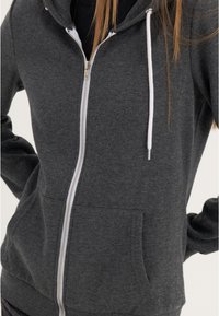 Sudadera gris con cremallera hecha de tela suave, con un bolsillo delantero, cremallera blanca y cordones en la capucha. Diseño simple.