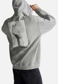 Persona che indossa un hoodie grigio con una grande grafica in bianco e nero di una mano che forma un cuore con le dita sul retro, abbinato a pantaloni scuri.