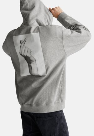 Persoon in een grijze hoodie met een groot zwart-wit grafisch ontwerp van een hand die een vingerhart maakt op de achterkant, gecombineerd met donkere broek.