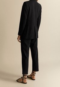 Musta räätälöity bleiseri, jossa on yksi takaintasku, yhdistetty mustiin kapeneviin housuihin ja mustiin remmissandaaleihin. Sujuva kankaan rakenne.